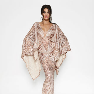 A model wearing a shimmering rose gold and beige sequined jumpsuit with dramatic draped cape sleeves, set against a plain white studio background.