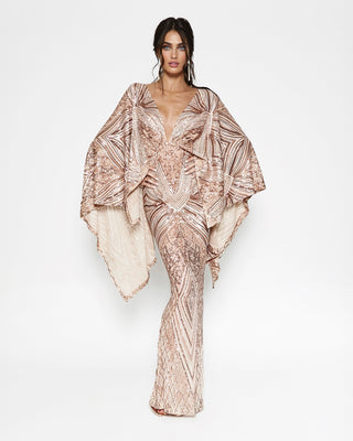 A model wearing a floor-length, deep V-neck gown with wide draped cape sleeves, featuring an intricate metallic rose gold and champagne patterned print, set against a plain white studio background.